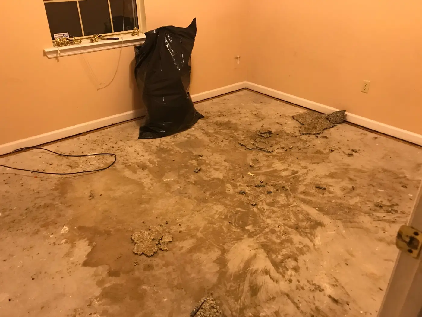 During: the same room with carpet pulled up, exposed concrete subfloor, and a bag of debris in the corner