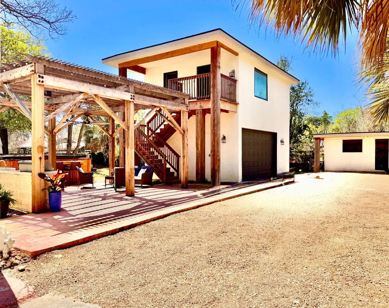 After: a finished two-story white stucco garage and guest apartment with a red-brown metal roof, a wood pergola, an exterior staircase, and a hot tub and outdoor kitchen underneath