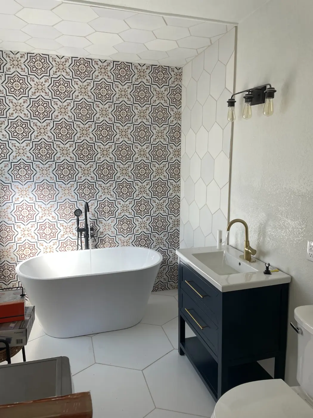 The finished guest bathroom with a patterned cement-tile accent wall, a freestanding soaking tub, a navy vanity, and brass fixtures