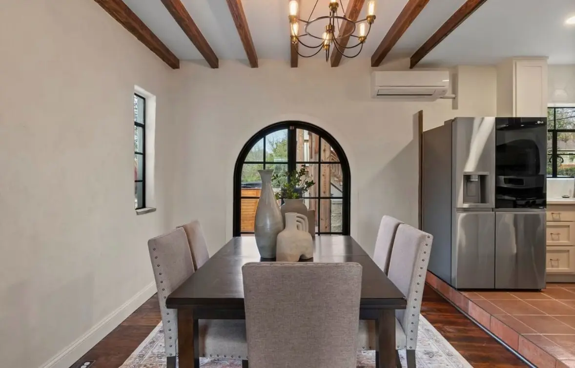 After: a dining room with a dark wood table, an arched black-framed exterior door, wood beams, and a chandelier