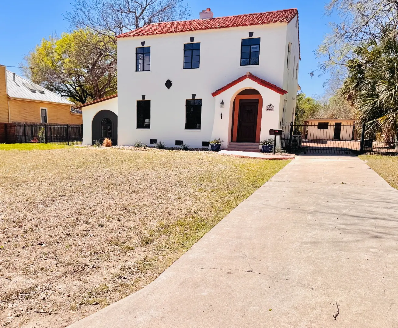 After: the same house with a red tile roof, crisp white stucco, a lush green lawn, and a black iron fence and gate