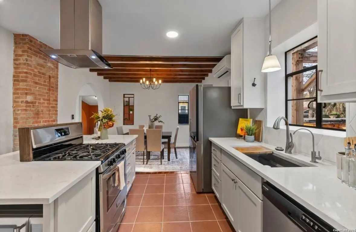 After: the finished kitchen with the original cabinets painted white, the counter behind the stove cut down, quartz counters, the cleaned-up brick chimney as a feature wall, terracotta tile floor, and a beamed ceiling
