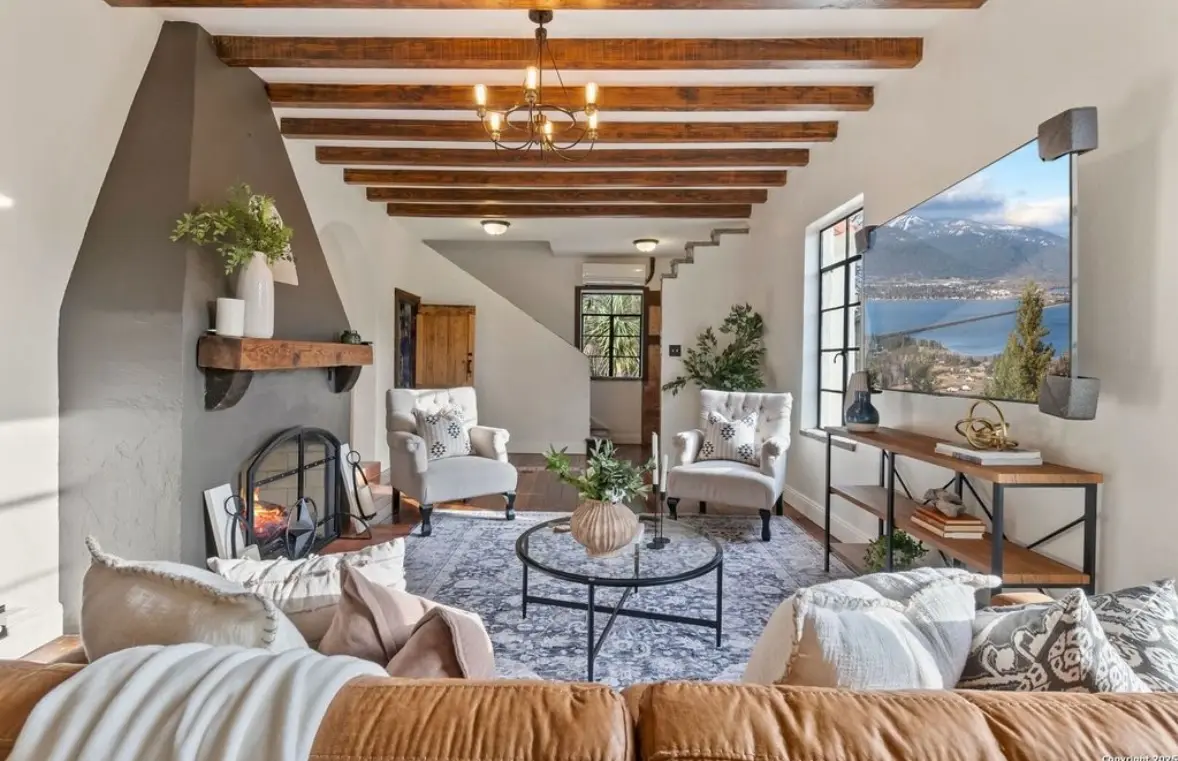 After: the finished living room with a beamed ceiling, chandelier, stone fireplace, cream sectional, and wood stairs