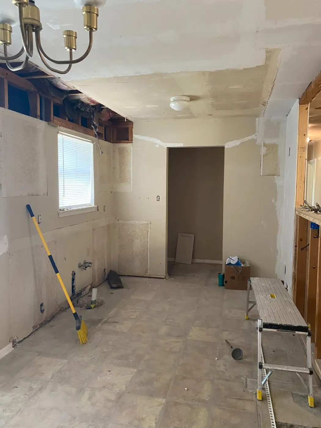 A gutted kitchen mid-demo, with the popcorn ceiling scraped away, cabinets removed, and a broom leaning against the stripped wall