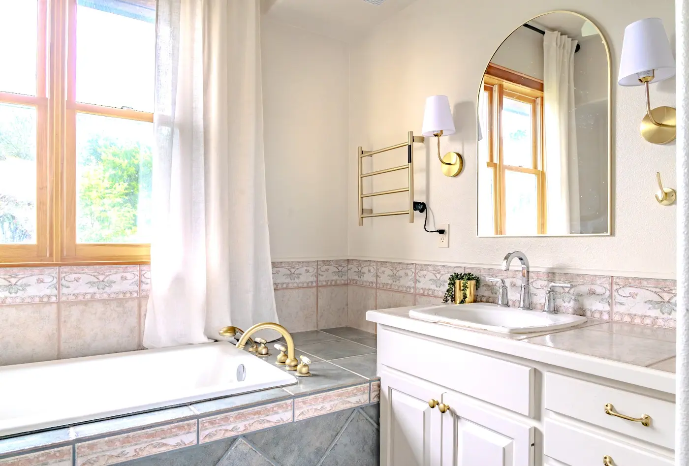 A close-up of the finished primary bath vanity with brass fixtures, a brass towel warmer, and an arched mirror