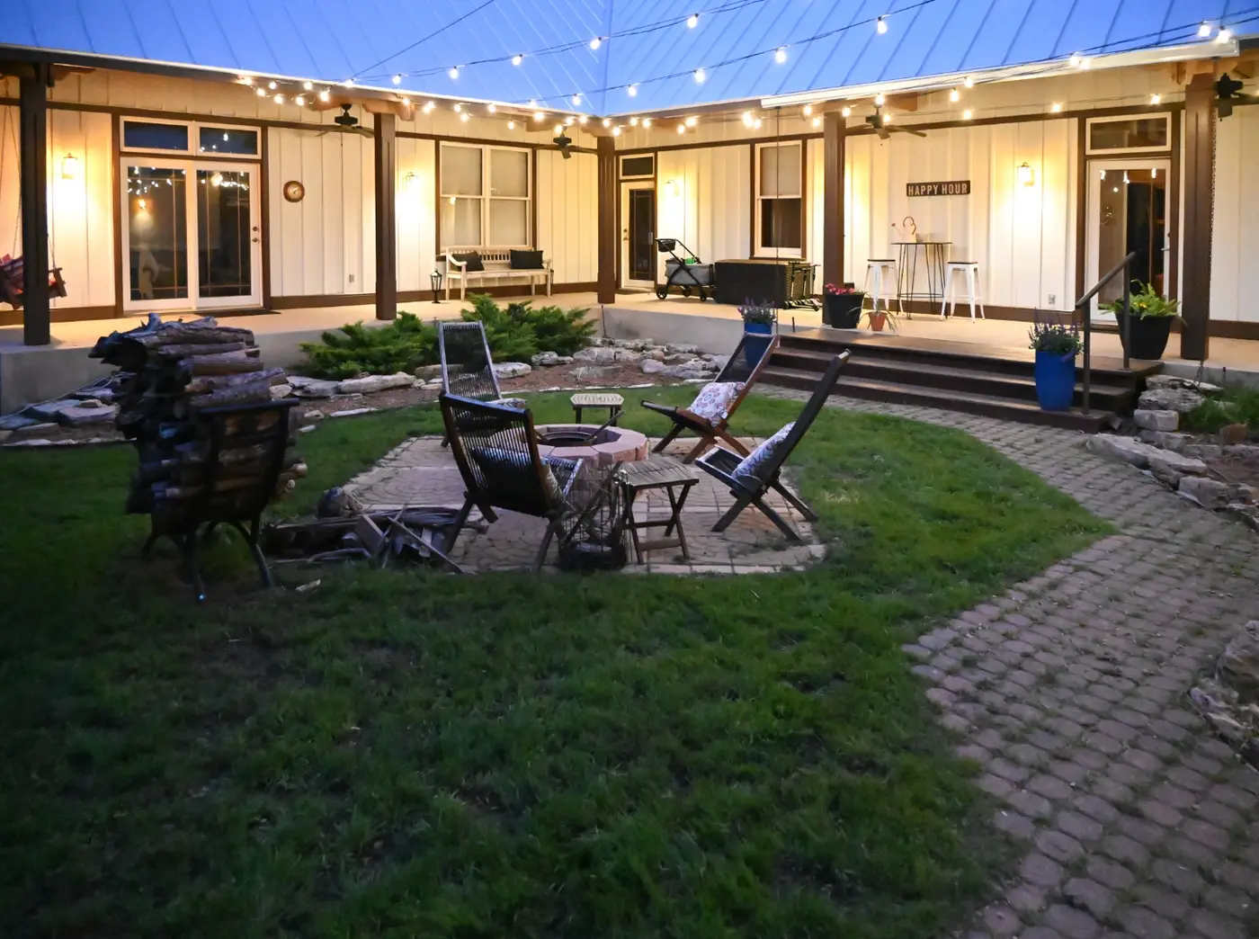 The same courtyard at twilight with string lights overhead, a fire pit and lounge chairs on the lawn, and a Happy Hour sign on the porch