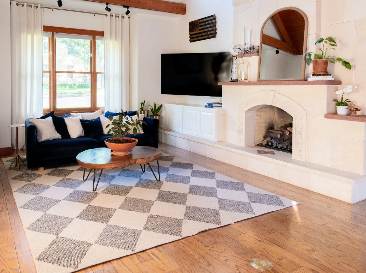 After: the same fireplace now cream-colored with a mirror over the mantel, anchoring a living room with a navy sofa, checkered rug, and mounted TV