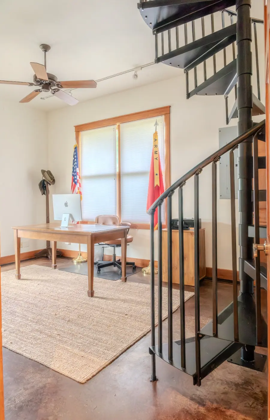 After: the same loft furnished as a home office with a wood desk, an iMac, an American flag, and an Army guidon flag beside the same spiral staircase